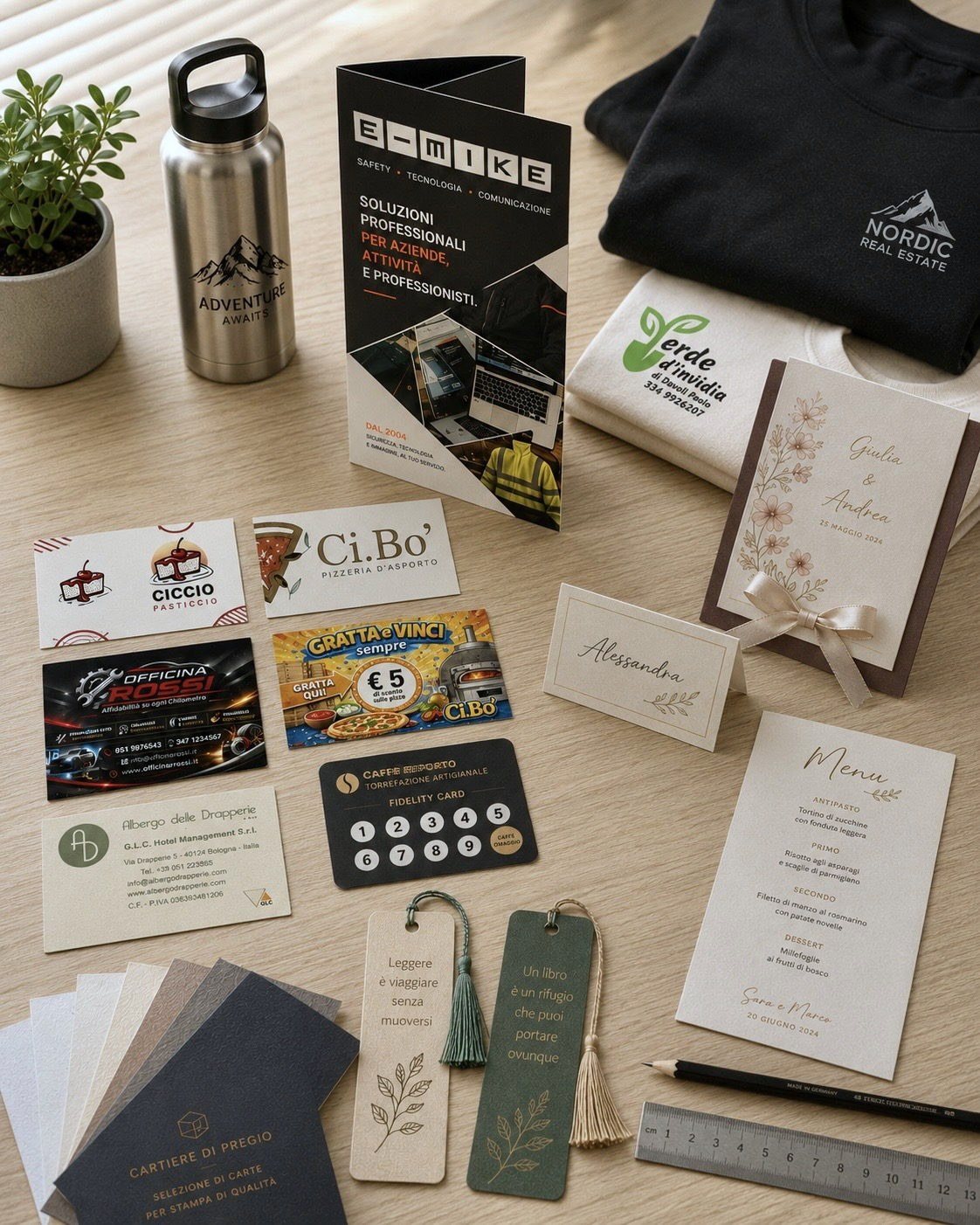 Lavori E-MIKE Studio su un piano in legno: felpa e t-shirt personalizzate, partecipazione di nozze con segnaposto e menu, biglietti da visita, depliant e segnalibri stampati, borraccia incisa al laser.
